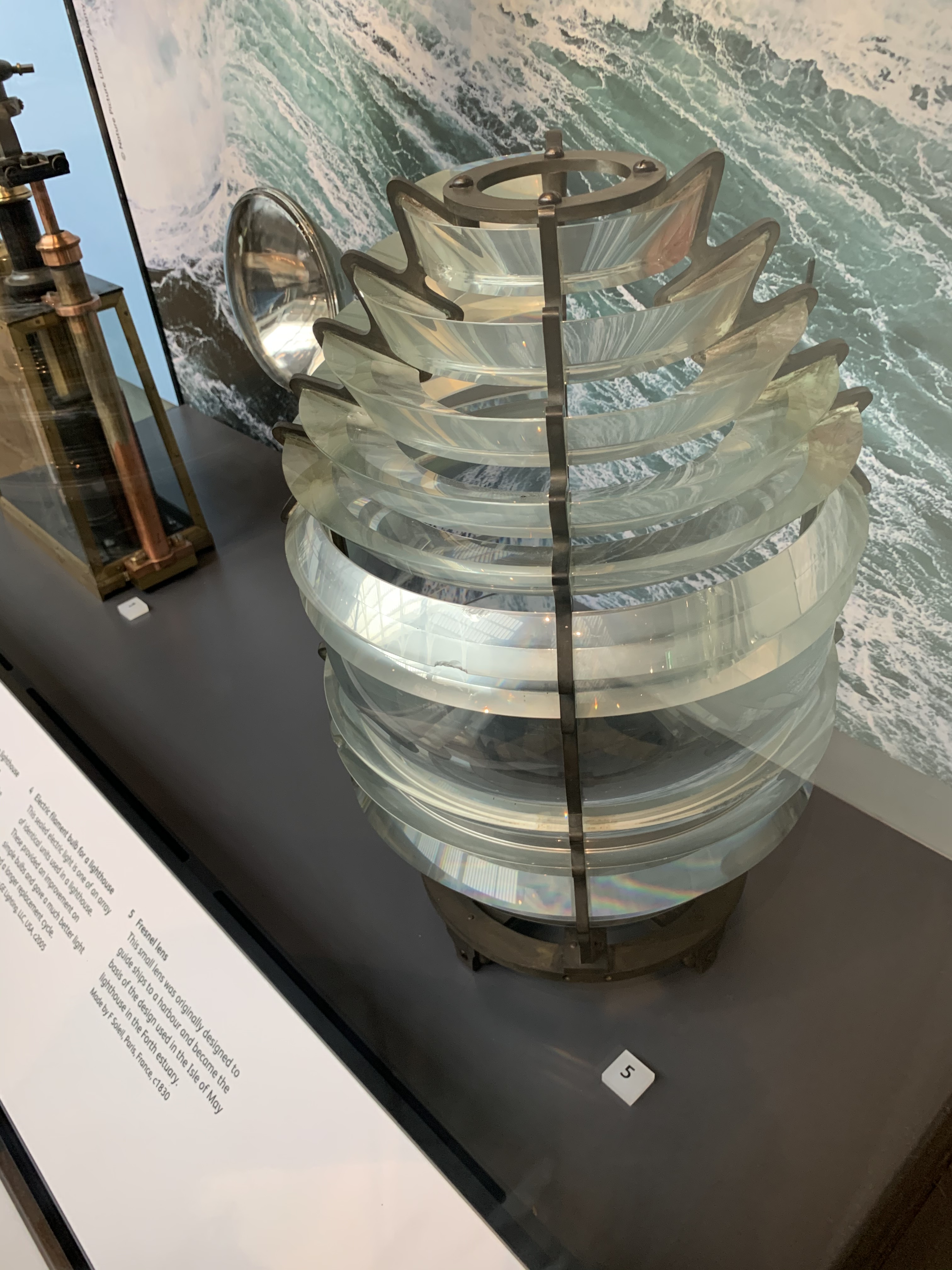 Unlit Fresnel lighthouse lens on museum display, with concentric glass sections clearly visible. Museum label visible in photograph identifies F. Soleil, Paris, 1830.